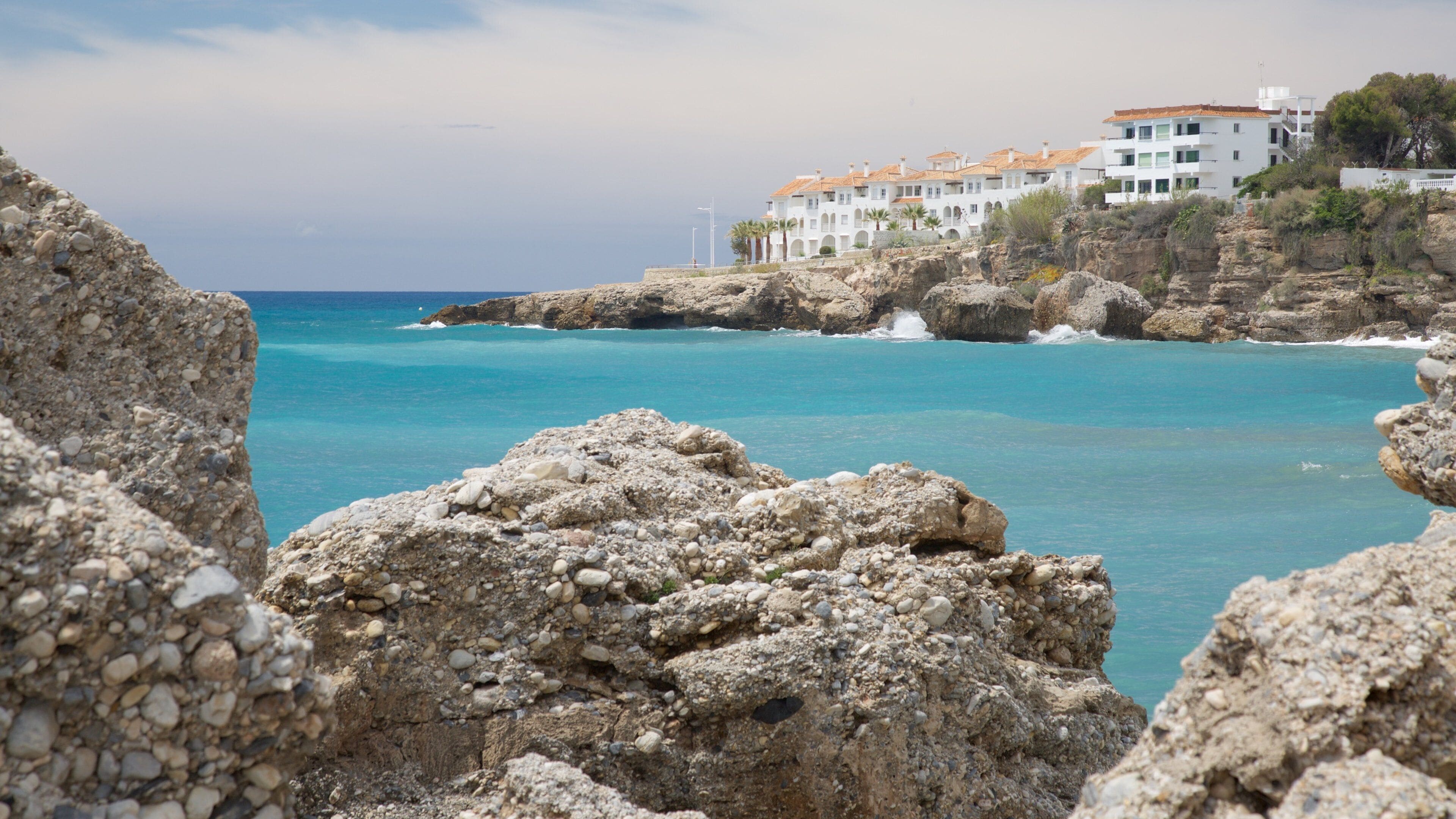 A coastal landscape with rocky terrain, clear turquoise waters, and white buildings with orange roofs on a hillside.