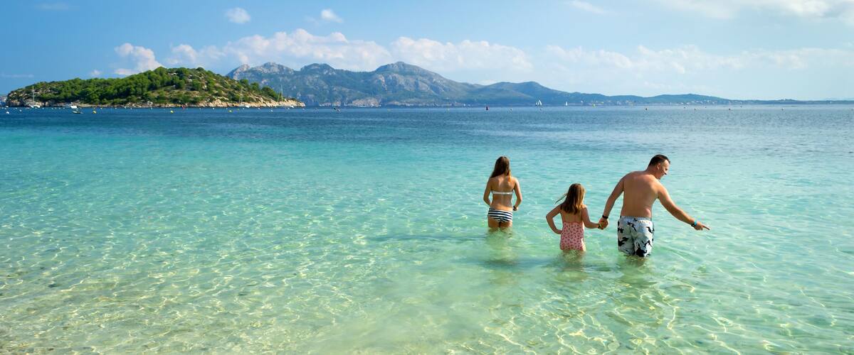 A family walking in clear turquoise waters with a mountainous island in the background.