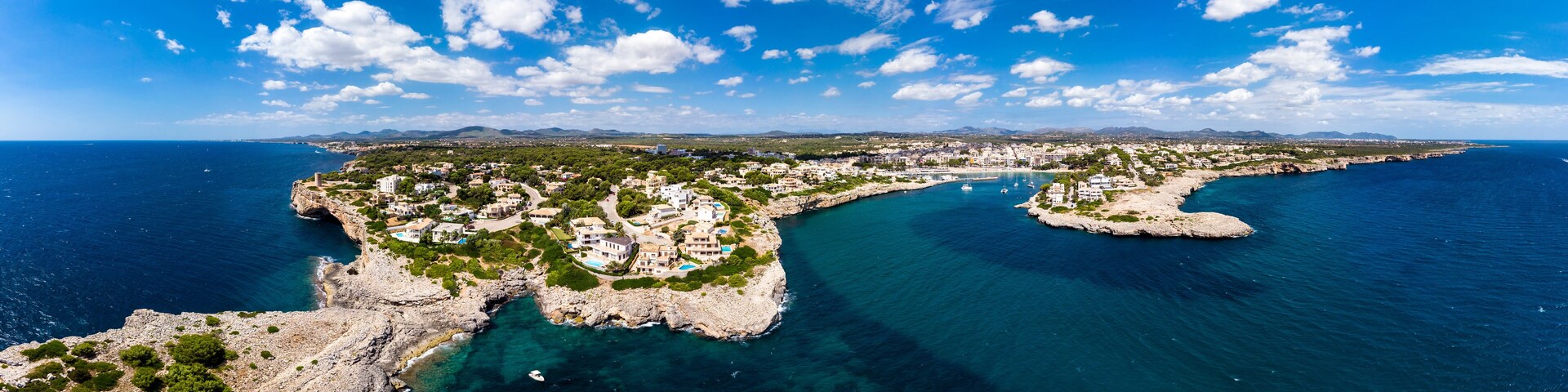 Un pueblo costero con un litoral rocoso, aguas azules cristalinas y montañas al fondo.