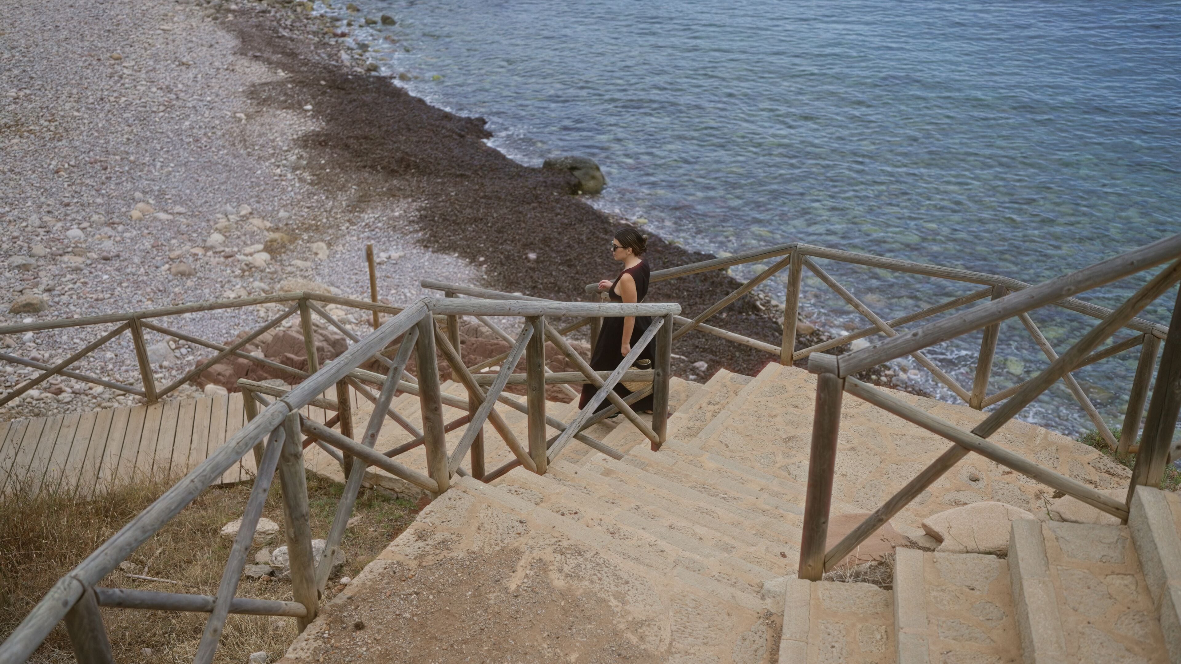 Young woman walking down wooden stairs by the rocky beach at port de valldemossa in mallorca, spain with the clear blue sea in the background