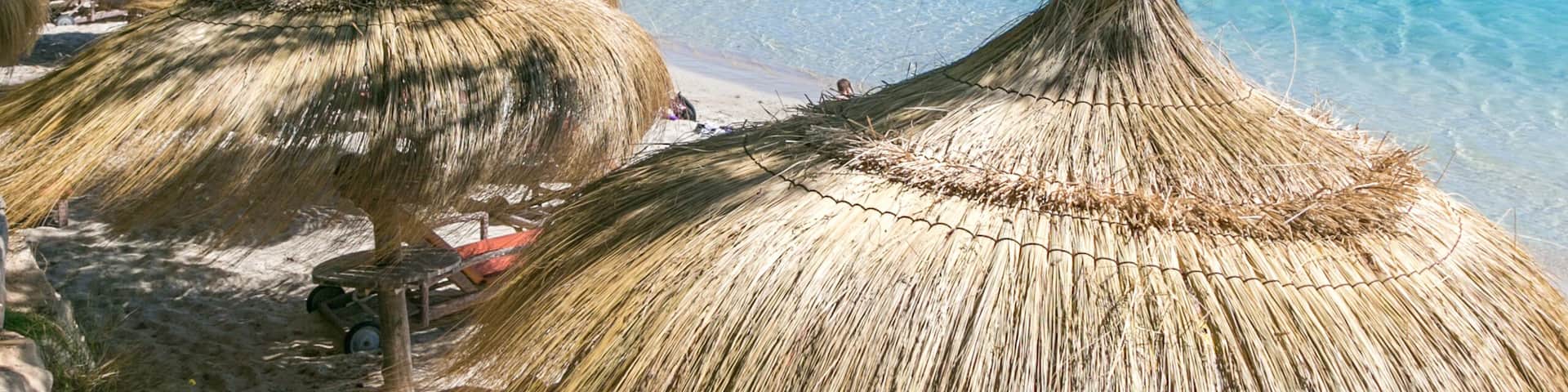 A beach with thatched umbrellas, clear turquoise water, and a mountainous backdrop.