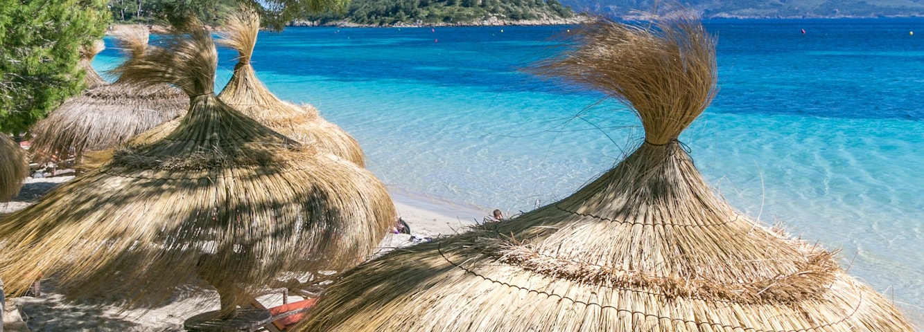 Una playa con sombrillas de paja, agua turquesa cristalina y montañas al fondo.