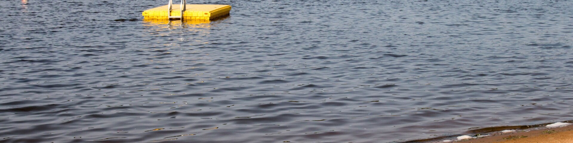 Una playa arenosa con aguas tranquilas, un muelle flotante amarillo y una costa con vegetación al fondo.
