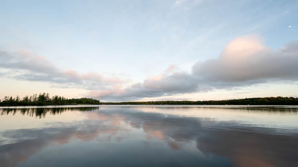 A calm lake with a clear reflection of the sky and trees, surrounded by a forested shoreline.