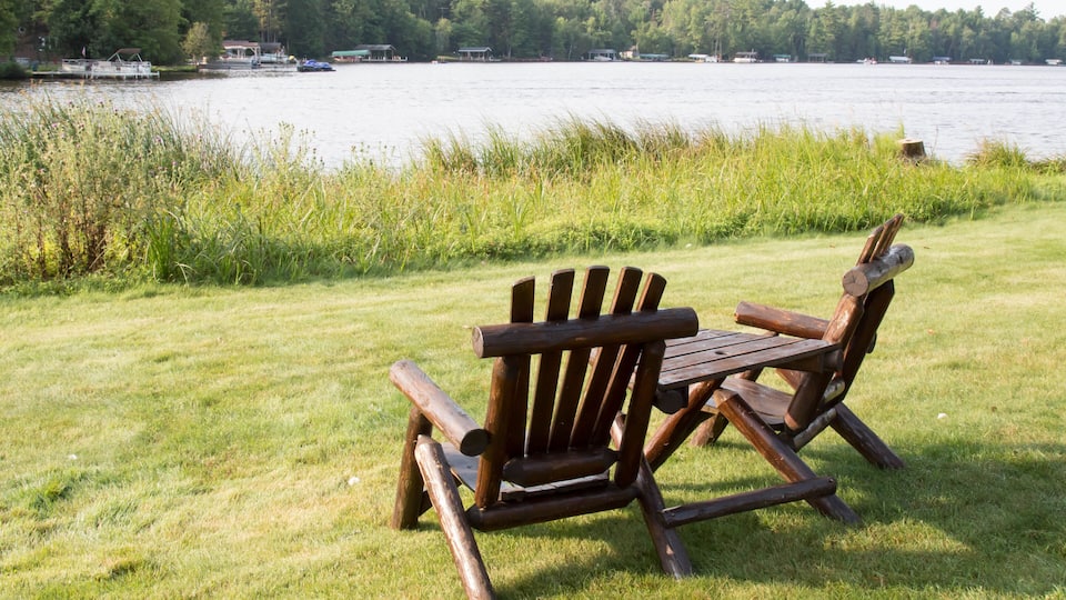Two Adirondack chairs on a grassy area by a lake with boats in the background.