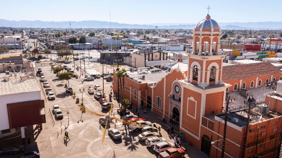 A cityscape with a prominent church featuring a dome and bell tower, surrounded by palm trees and a parking lot.
