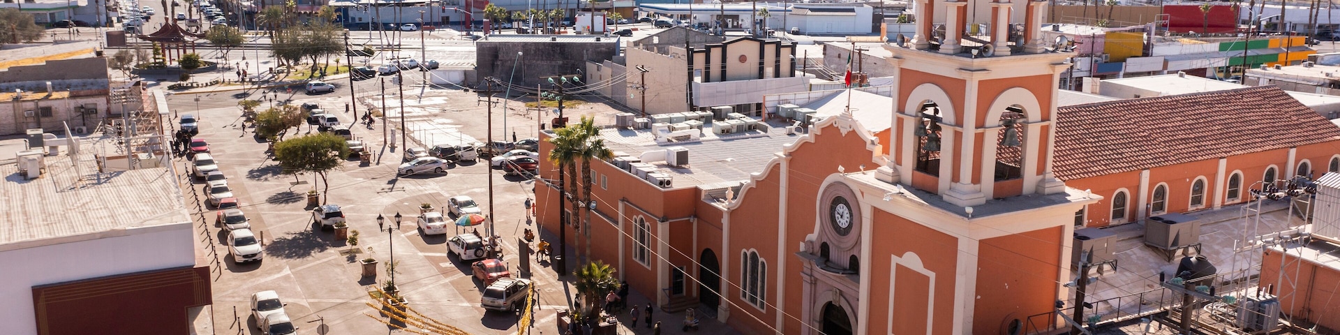 Un paisaje urbano con una iglesia destacada, que cuenta con una cúpula y un campanario, rodeada de palmeras y un estacionamiento.