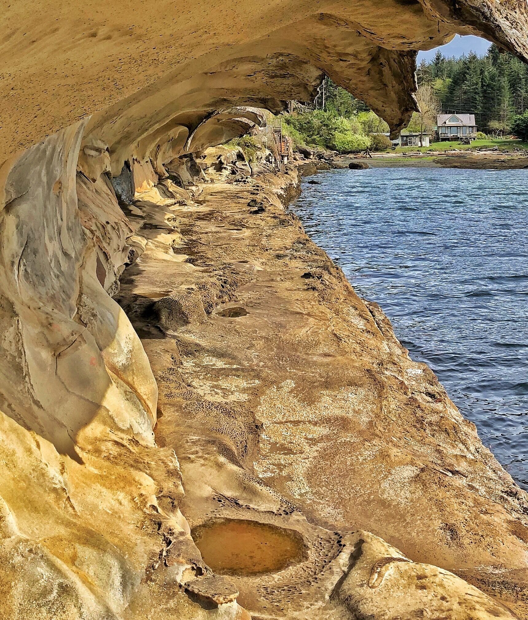 Uma costa rochosa com uma abertura de caverna, uma pequena construção e uma casa ao fundo.