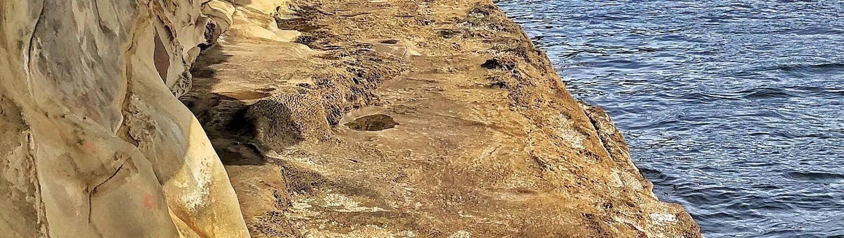 Uma costa rochosa com uma abertura de caverna, uma pequena construção e uma casa ao fundo.