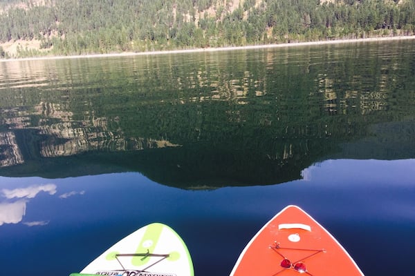 Ein ruhiger See mit Spiegelungen von Bäumen und einem bewaldeten Hang. Im Vordergrund sind zwei Stand-Up-Paddles zu sehen.