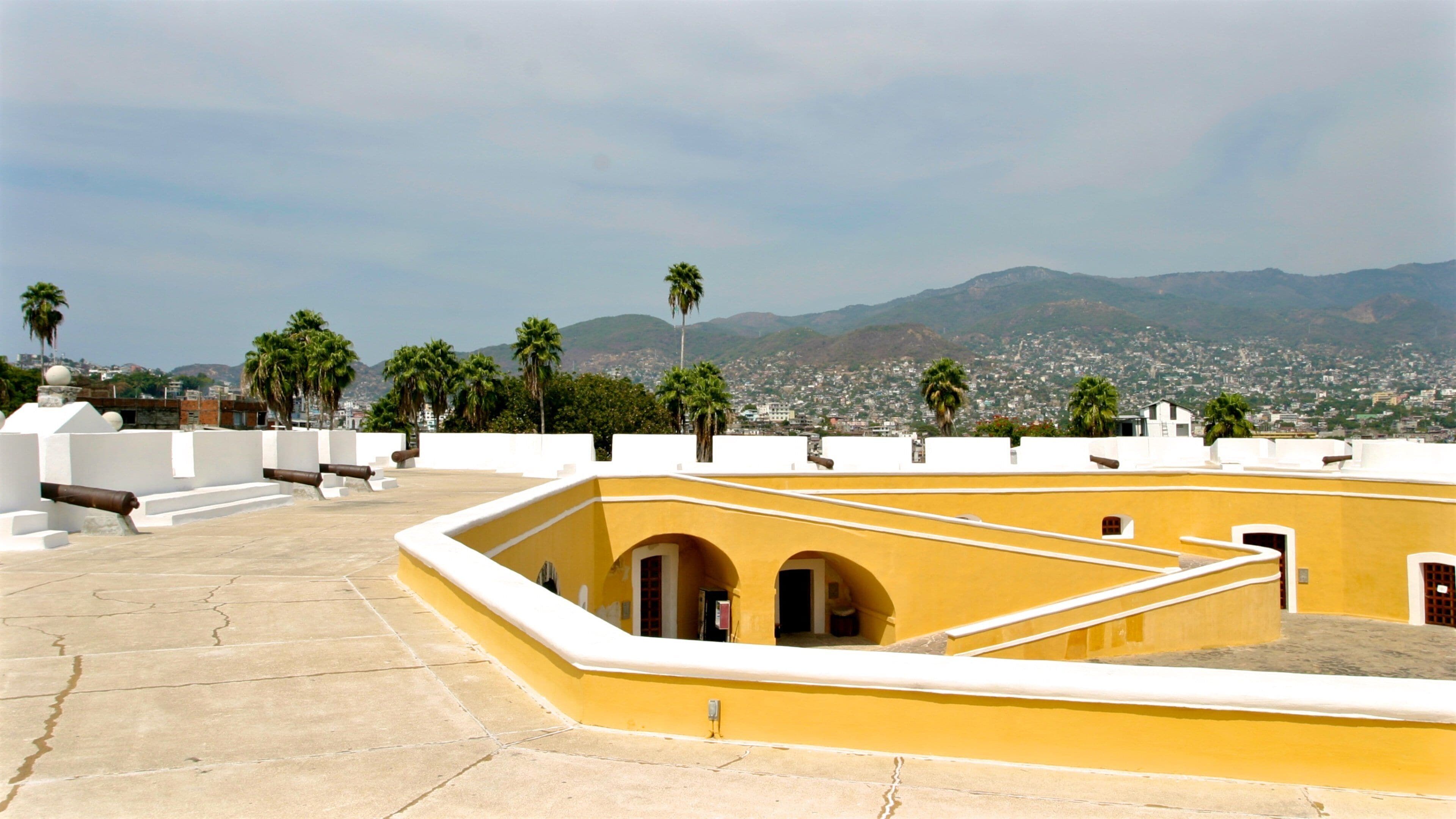 A yellow building with arched doorways, palm trees, and a cityscape in the background.