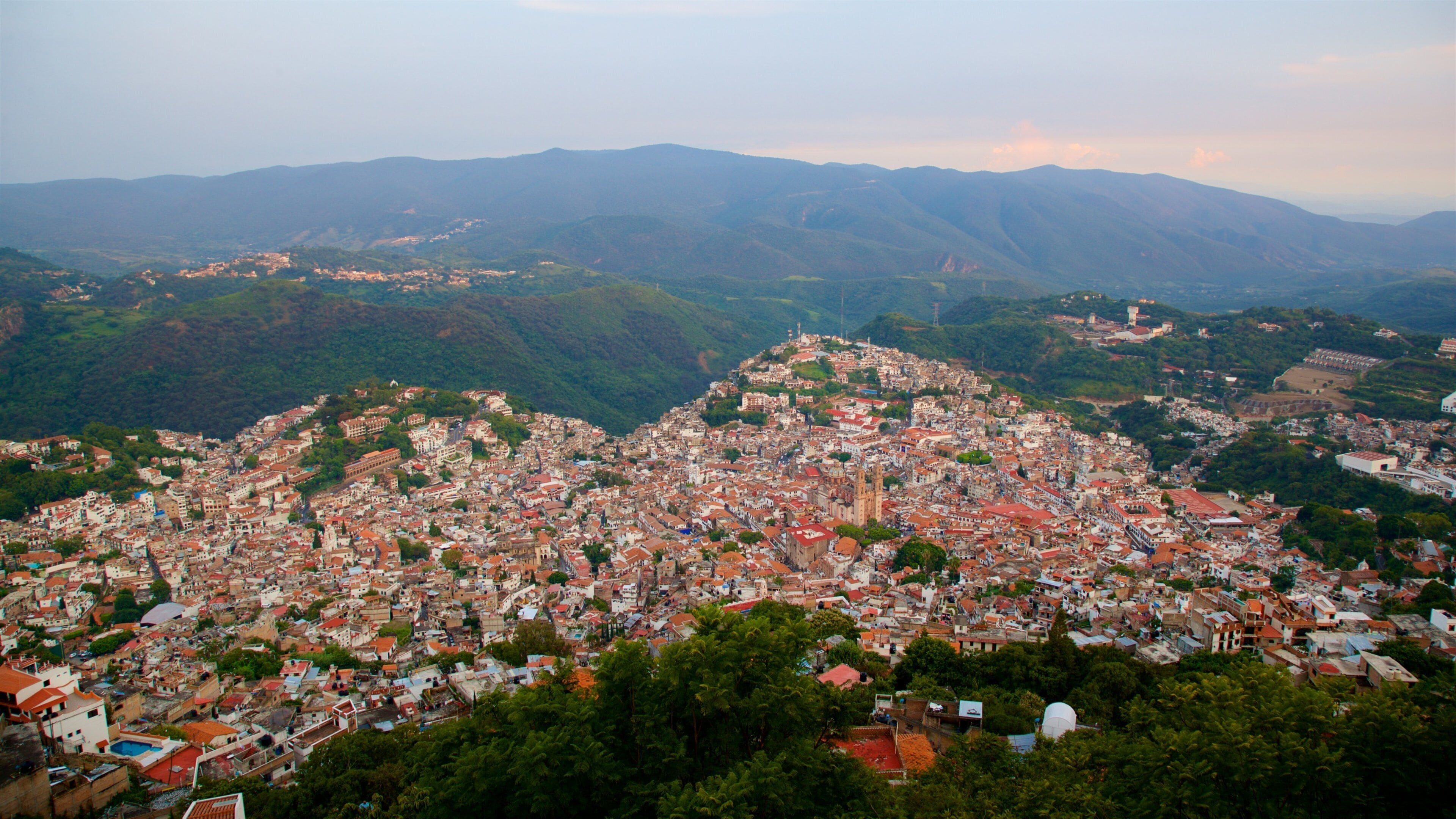 A densely packed city with red-roofed buildings, surrounded by lush green hills and mountains.