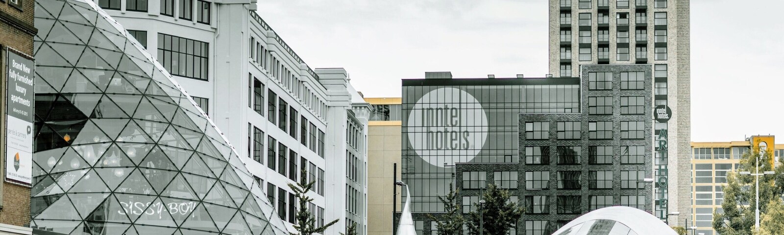 Una plaza urbana moderna con una estructura de cúpula de vidrio, rodeada de edificios contemporáneos y un rascacielos alto.