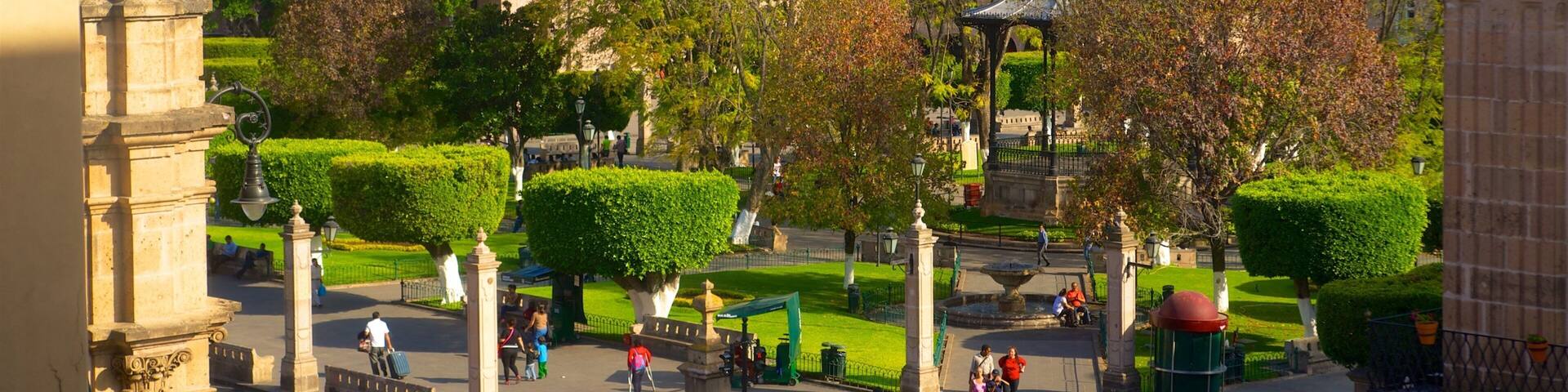 A park with a gazebo, neatly trimmed hedges, and a distant view of a hill with buildings.