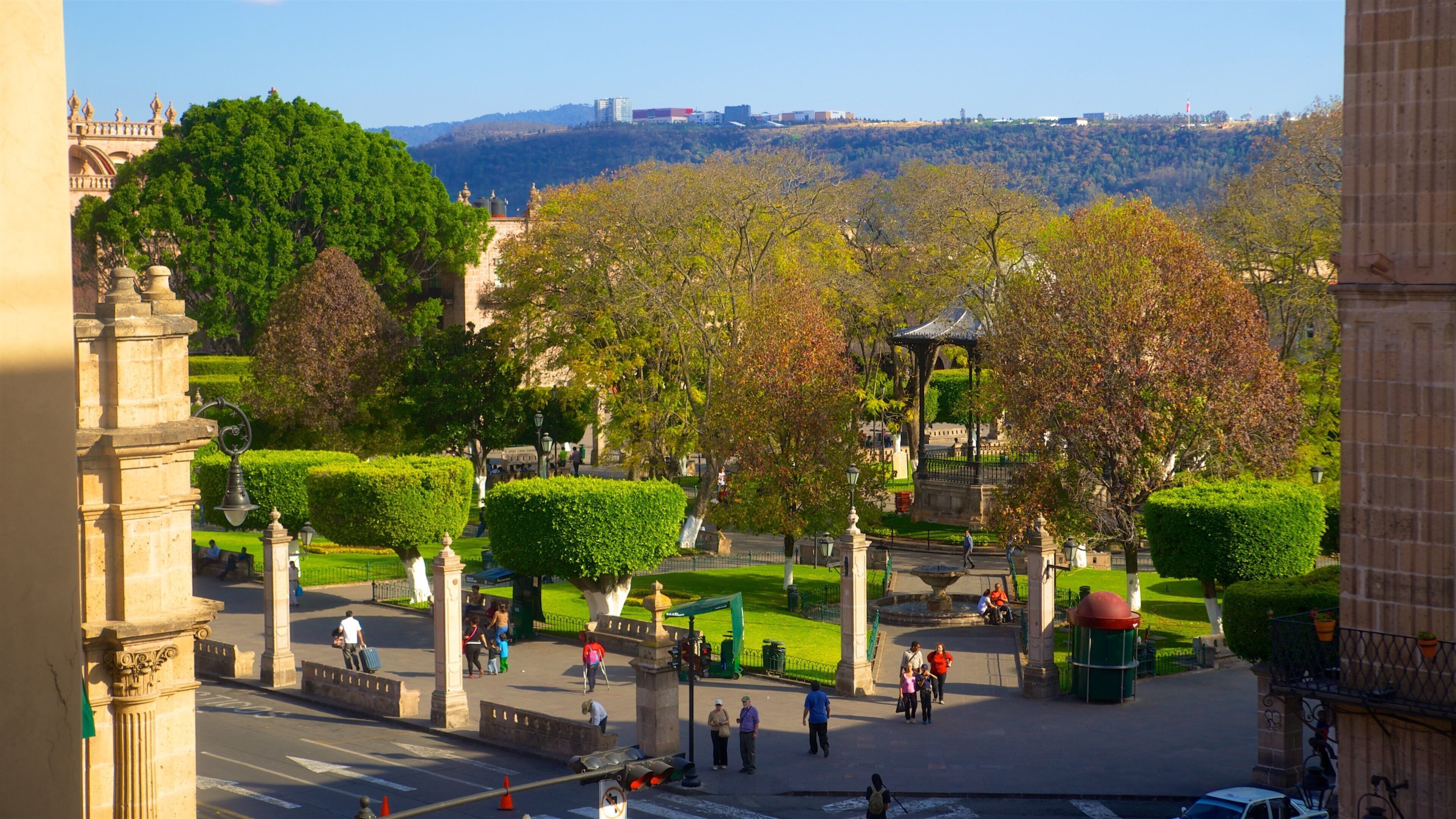 A park with a gazebo, neatly trimmed hedges, and a distant view of a hill with buildings.