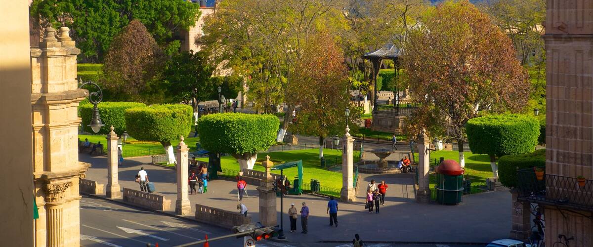 A park with a gazebo, neatly trimmed hedges, and a distant view of a hill with buildings.