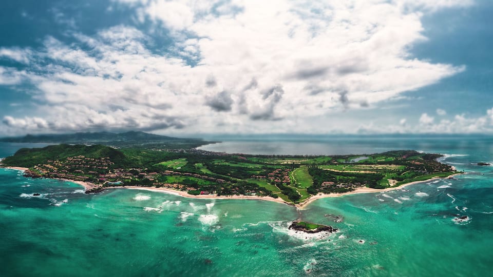 An aerial view of a tropical island with clear turquoise waters, a central peninsula, and a large cloud formation.