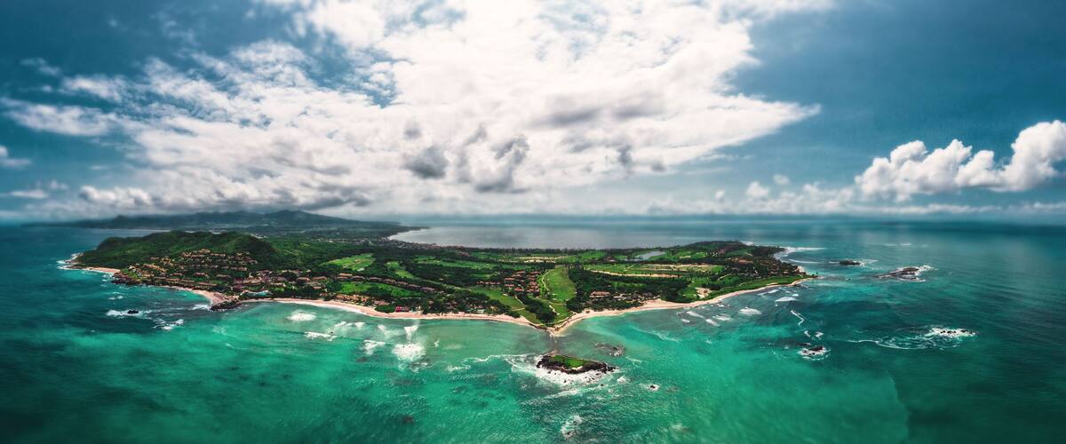 An aerial view of a tropical island with clear turquoise waters, a central peninsula, and a large cloud formation.