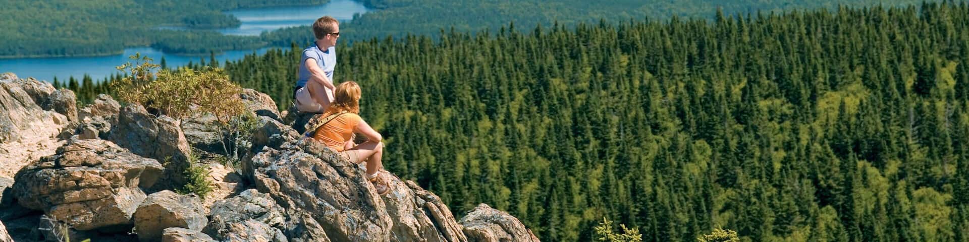 A person sitting on a rocky outcrop overlooking a forested valley with a lake.