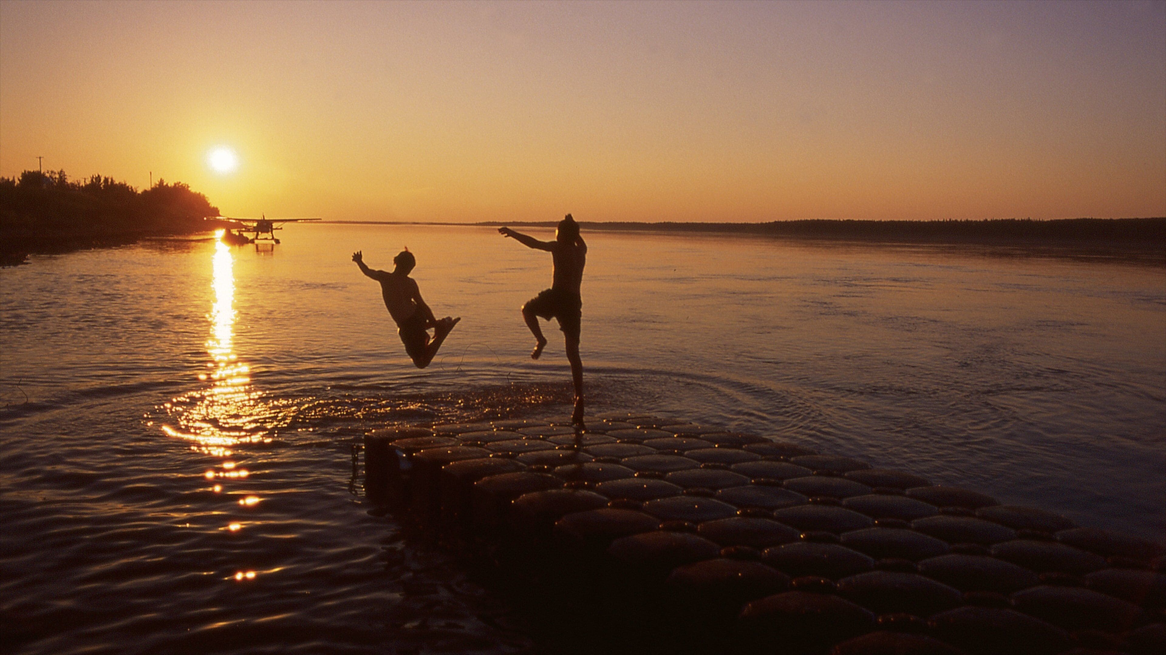 Duas pessoas saltam na água ao pôr do sol, com um barco ao fundo.
