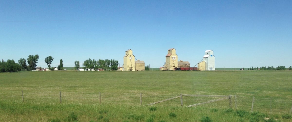 A rural landscape with a large open field, a fence, and three grain silos.