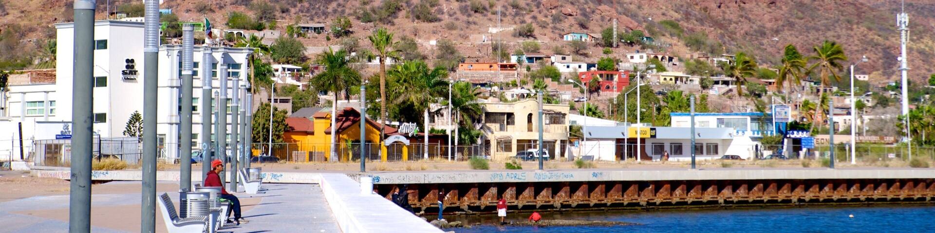 A coastal town with a prominent rocky mountain in the background, a waterfront promenade, and residential buildings.