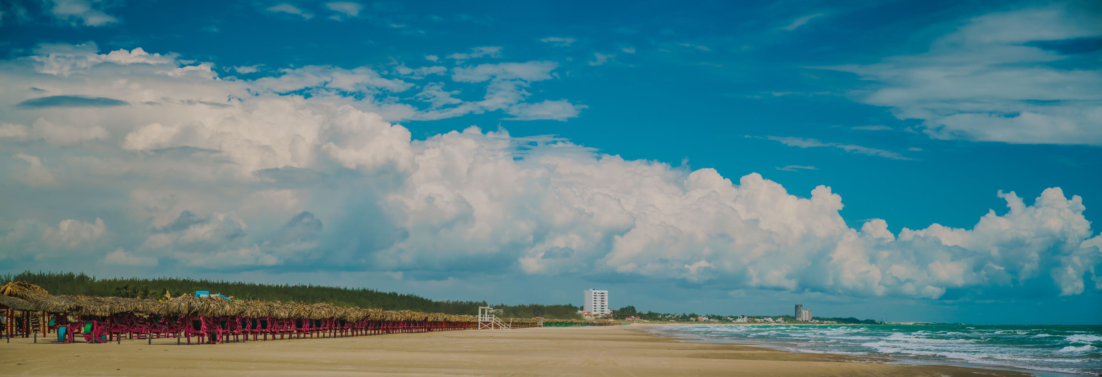 A beach with a thatched-roof structure, a lifeguard tower, and a distant building.