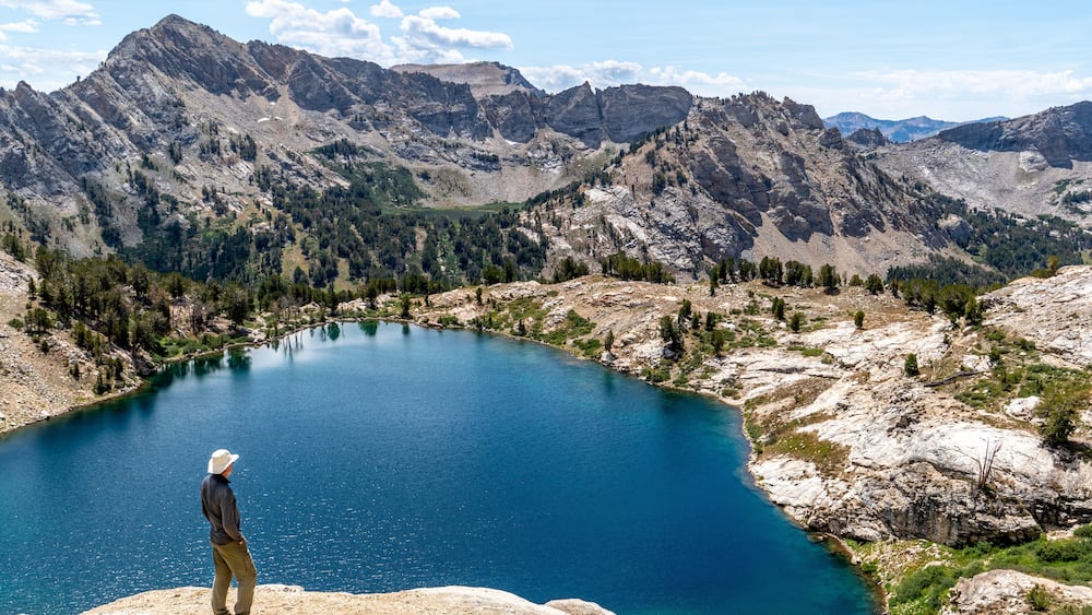 Man, hiker, standing, watching a scene of an alpine lake and rugged mountains with trees, rocks, and puffy clouds in the blue sky, Liberty Lake, Ruby Mountain Range, Elko, Nevada