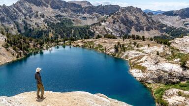 Man, hiker, standing, watching a scene of an alpine lake and rugged mountains with trees, rocks, and puffy clouds in the blue sky, Liberty Lake, Ruby Mountain Range, Elko, Nevada
