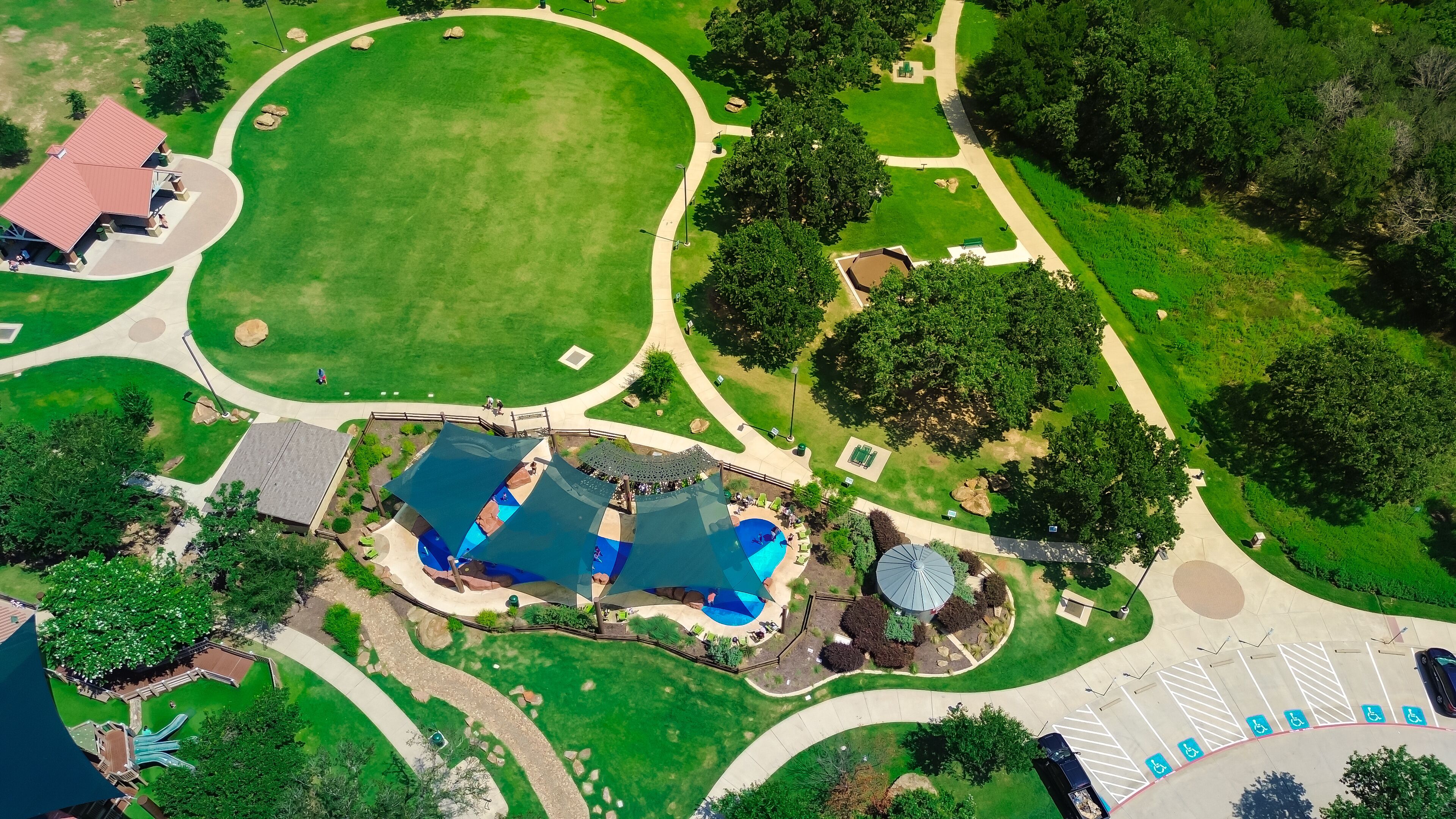 Suburban park with pavilion shelter, slash pad sail shade structure and diverse kids playing water in large modern children recreation center, lush green trees in Flower Mound, Texas, aerial view