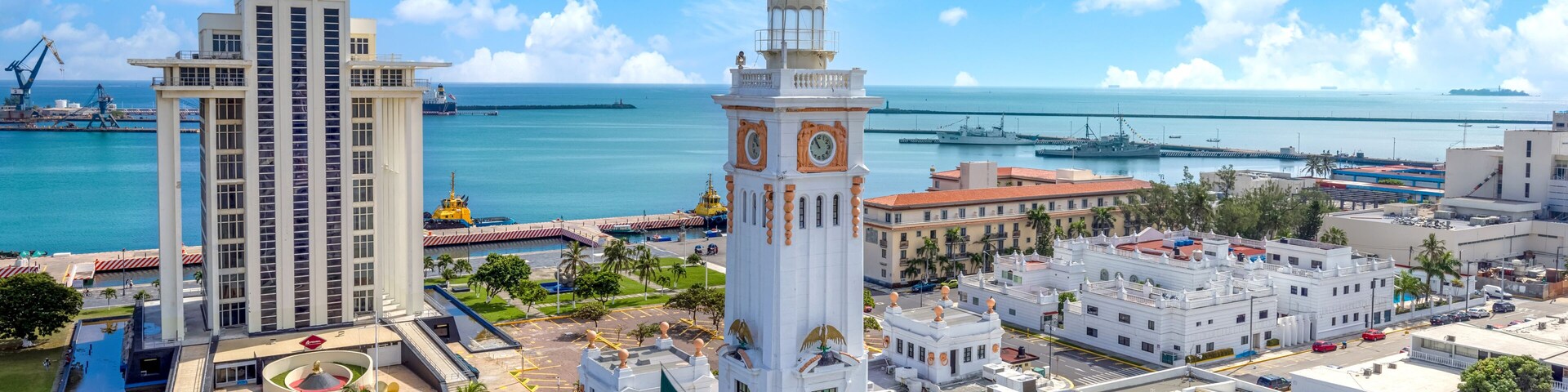 Un paisaje urbano costero con una imponente torre del reloj, un edificio alto y un puerto deportivo con barcos.