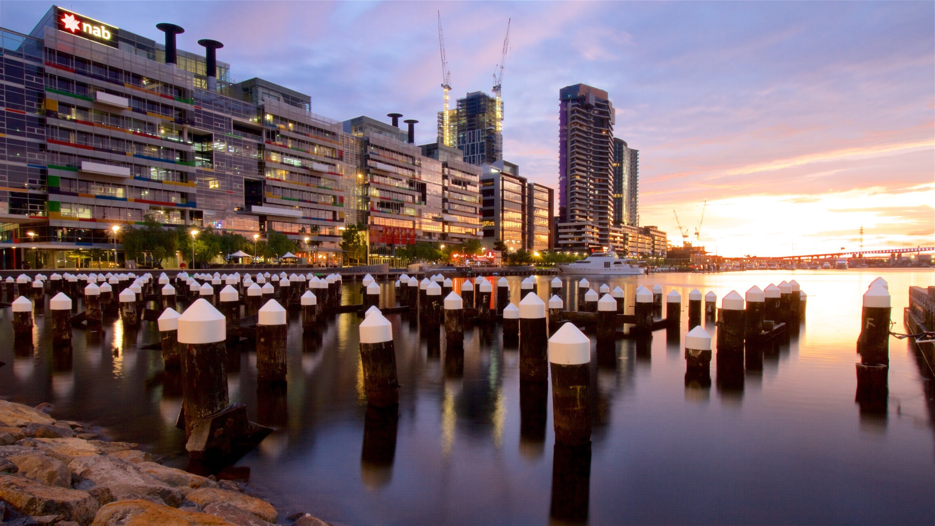 A waterfront cityscape with modern buildings, a marina with moored boats, and a sunset over the water.