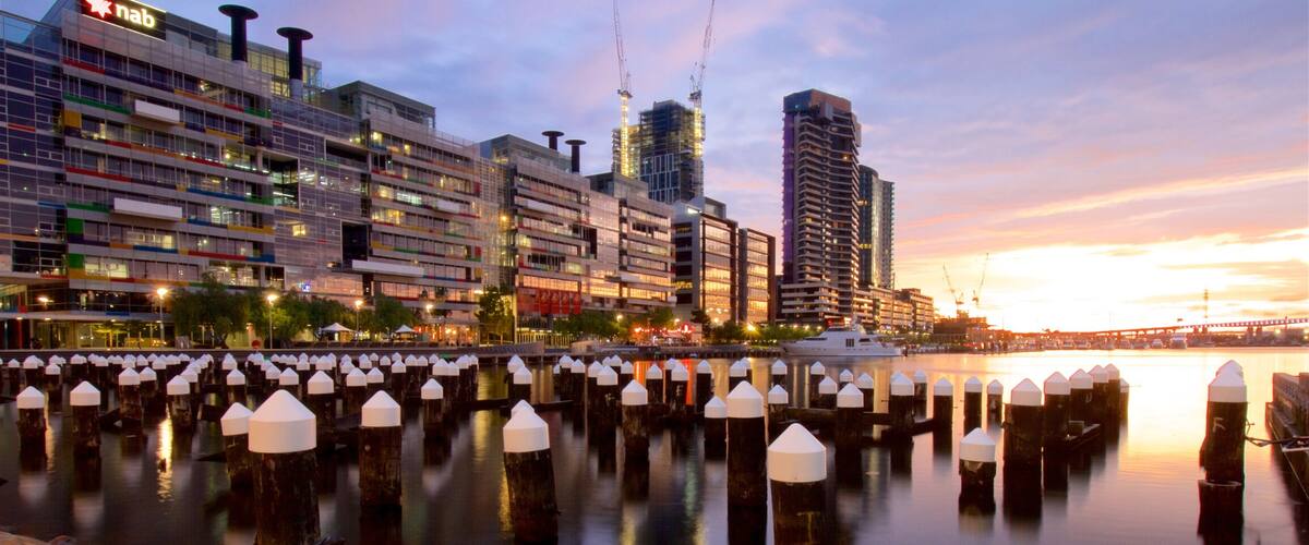 A waterfront cityscape with modern buildings, a marina with moored boats, and a sunset over the water.