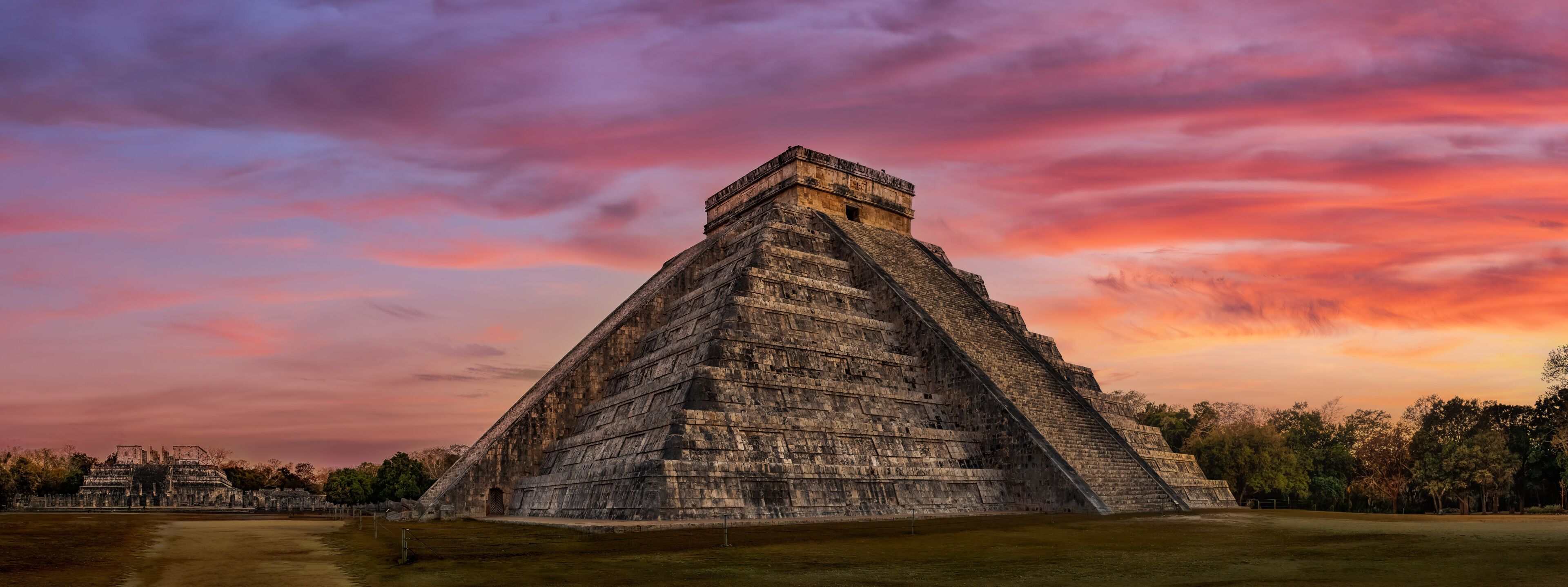 A large pyramid structure with a flat top, surrounded by a grassy area and trees, under a colorful sunset sky.