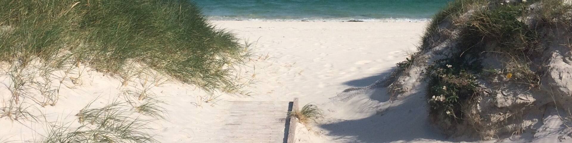 A sandy beach with a wooden boardwalk leading to the ocean, surrounded by grassy dunes and distant hills.