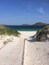 A sandy beach with a wooden boardwalk leading to the ocean, surrounded by grassy dunes and distant hills.