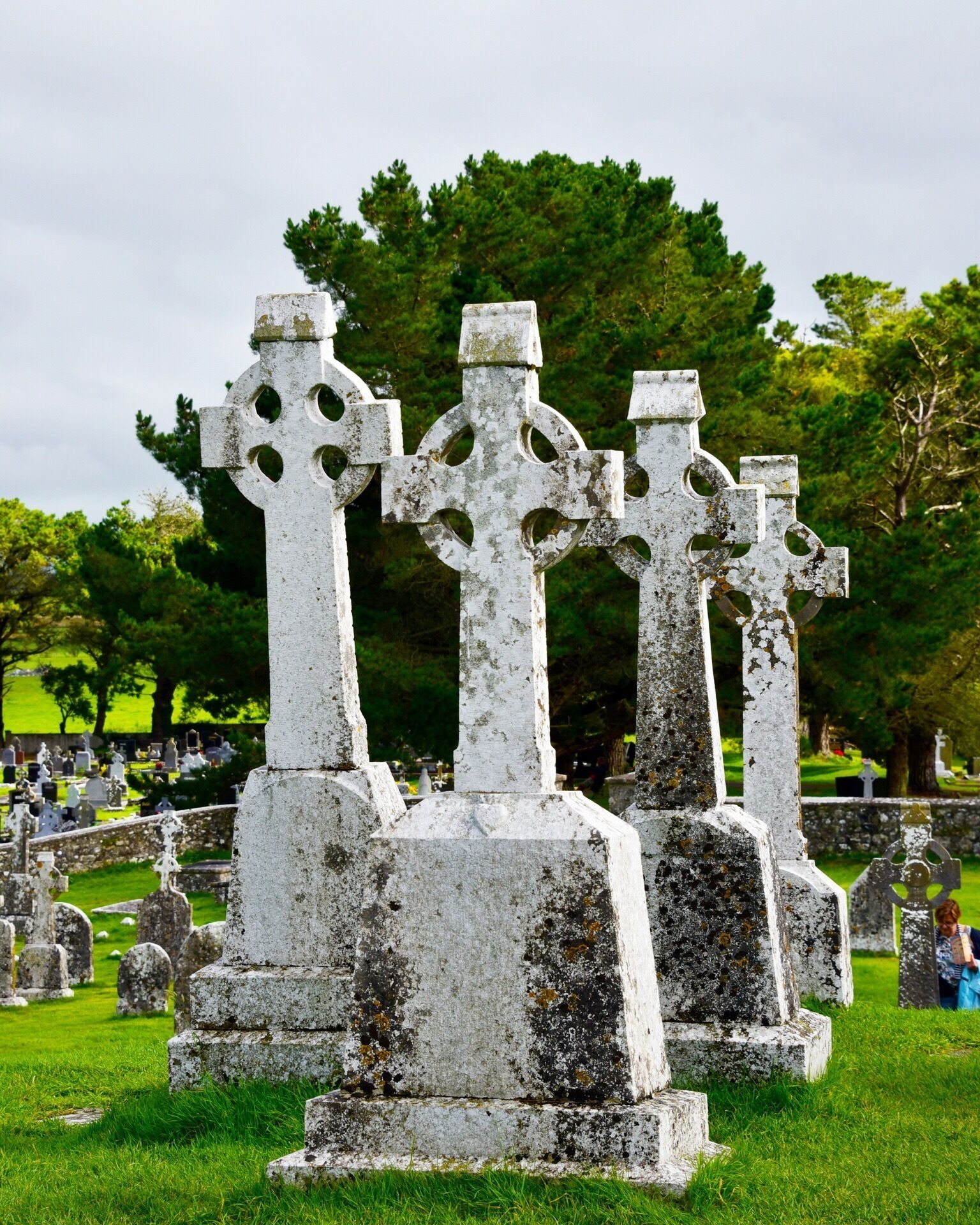 Un cementerio con antiguas cruces de piedra y frondosos árboles verdes al fondo.