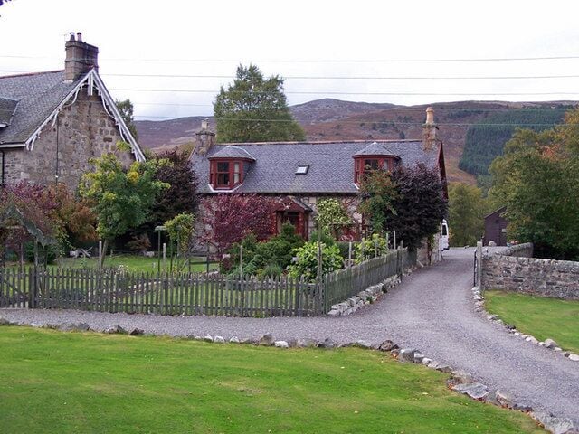 A rural stone house with a gravel driveway, surrounded by greenery and hills in the background.