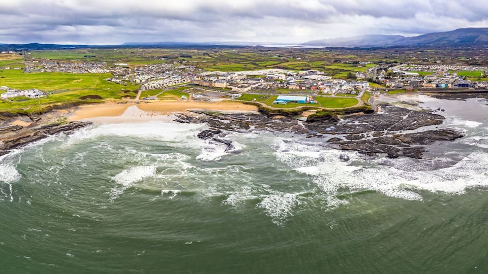 An aerial view of a coastal town with a sandy beach, rocky shoreline, and a mountainous backdrop.