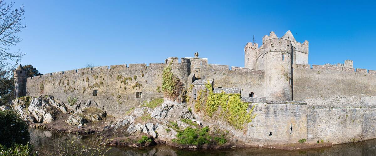 A historic stone castle with a moat, surrounded by rocky terrain and greenery.