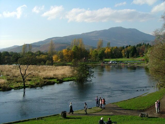 Un río con gente caminando por las orillas, una zona verde y montañas al fondo.
