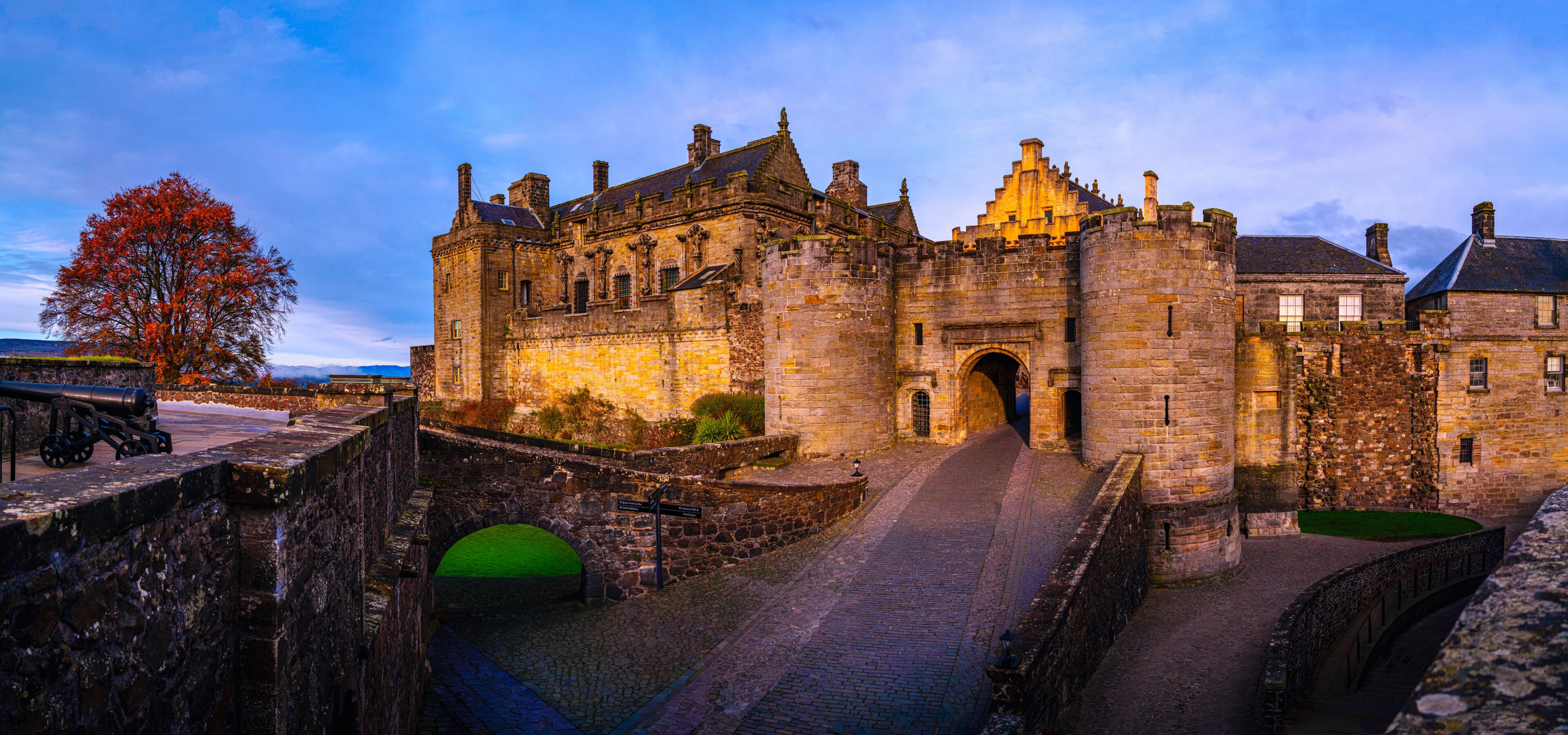 A historic stone castle with a large archway entrance, a cobblestone pathway, and a tree with autumn leaves.