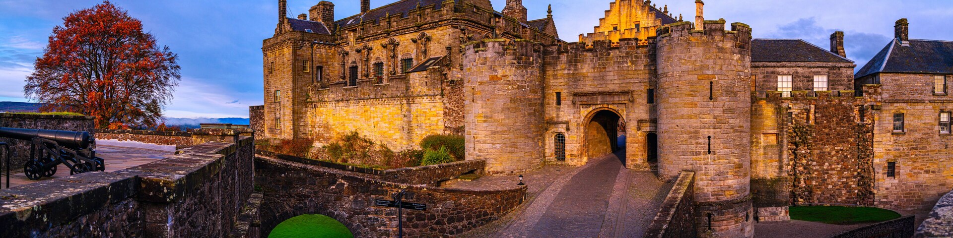 A historic stone castle with a large archway entrance, a cobblestone pathway, and a tree with autumn leaves.