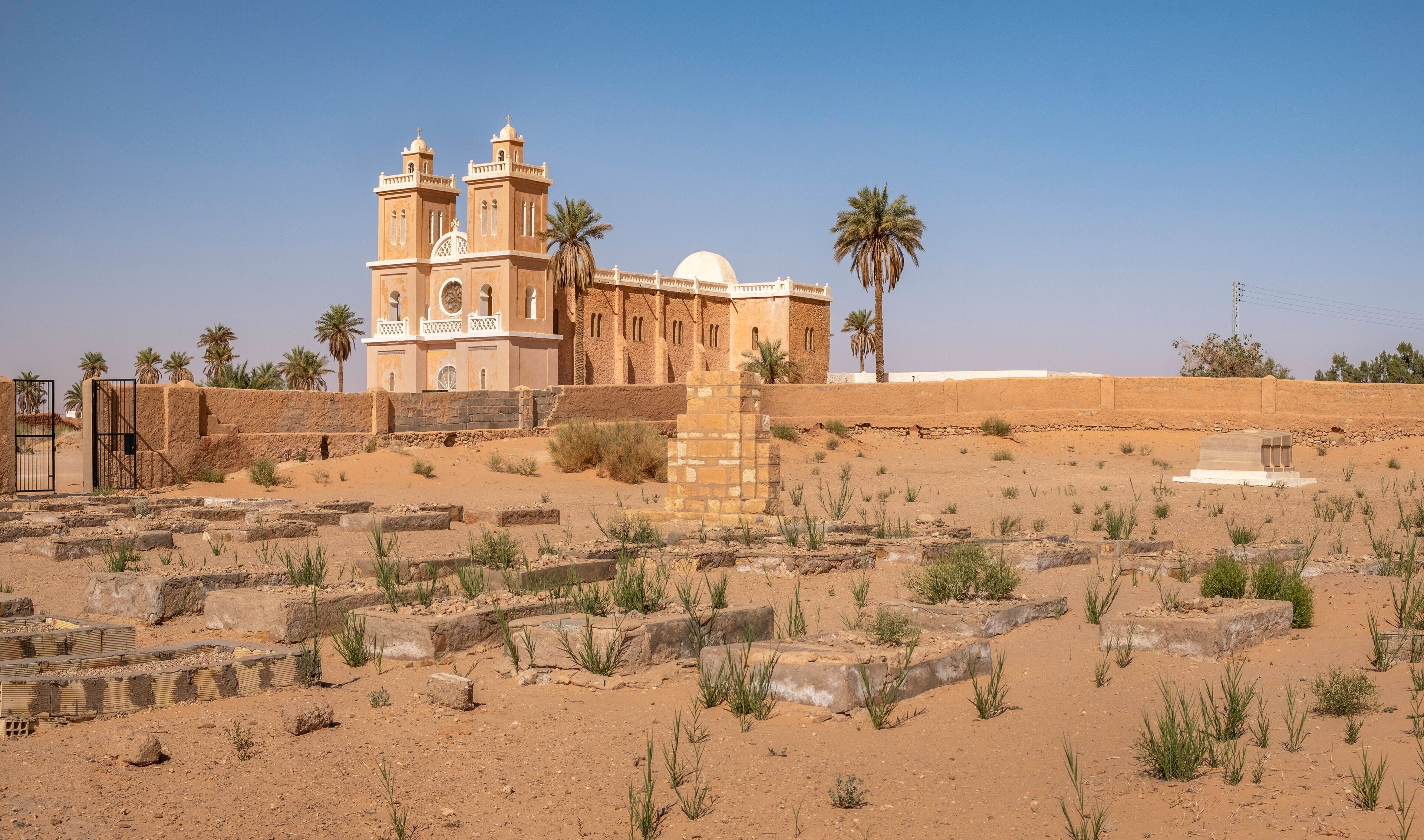 An ancient church with two towers and a dome, surrounded by desert ruins and palm trees.