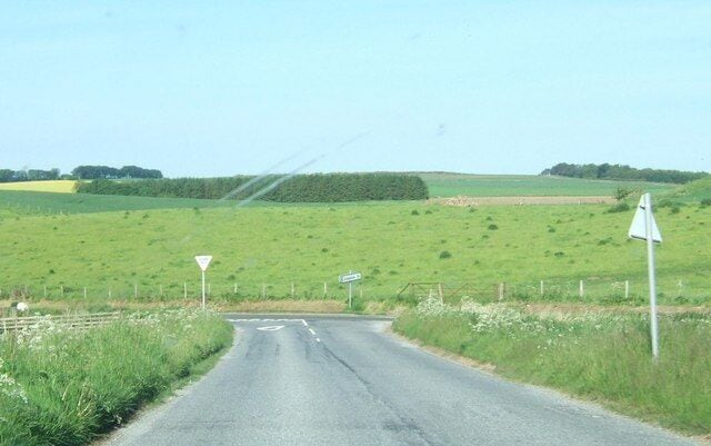 A rural road with a yield sign, surrounded by green fields and a clear sky.