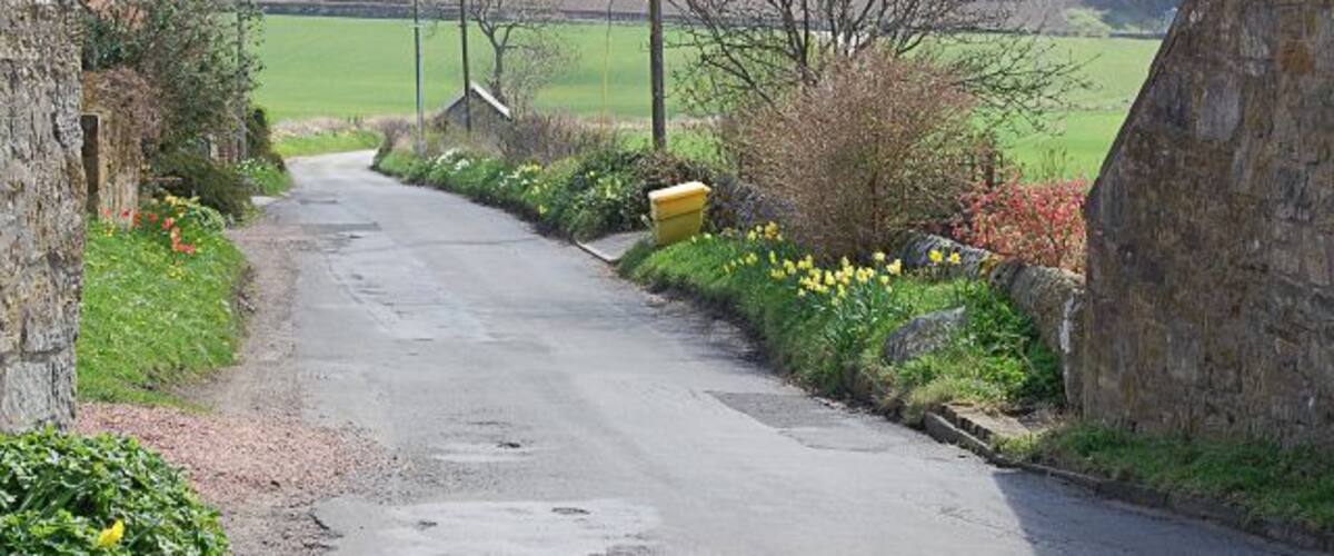 A narrow rural road flanked by stone walls and flowering plants, leading through a hilly landscape.