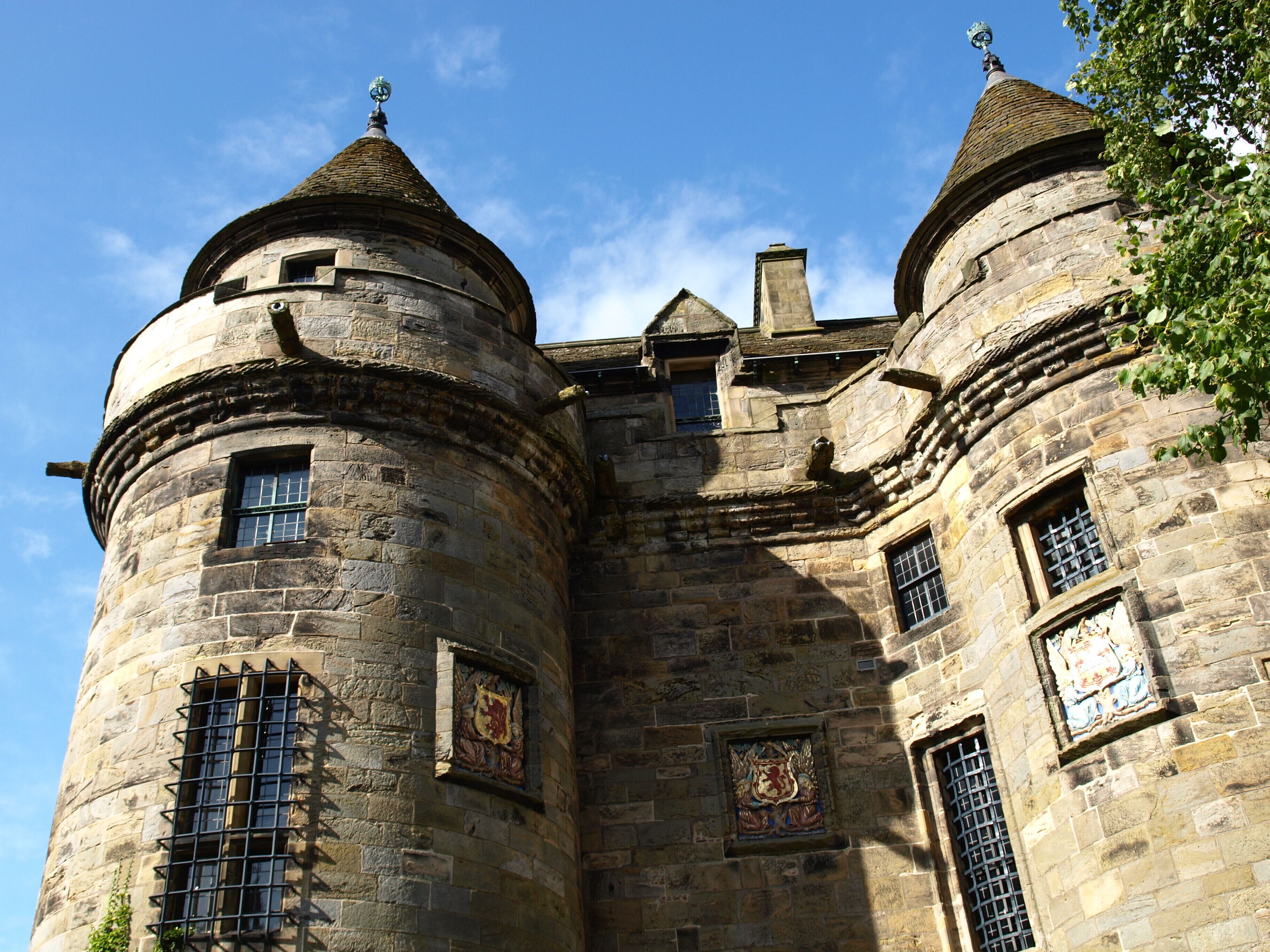 A historic stone castle with round towers, arched windows, and heraldic shields.
