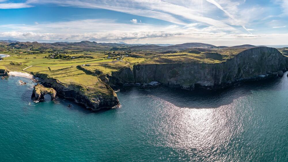 Un paysage côtier caractérisé par une falaise imposante, une petite île surmontée d’une arche rocheuse et, au loin, une chaîne de montagnes.