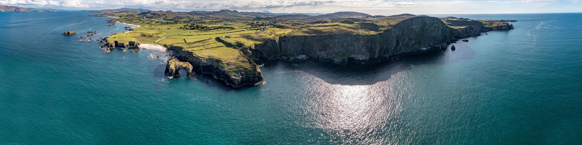 A coastal landscape with a prominent cliff, a small island with a rock arch, and a distant mountain range.