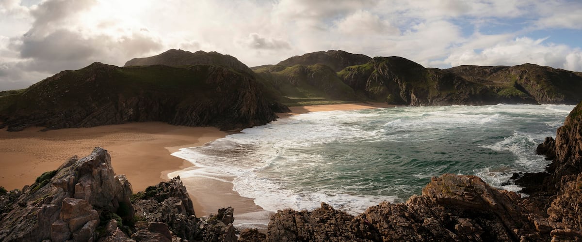A rugged coastline with rocky cliffs, a sandy beach, and a turbulent sea.